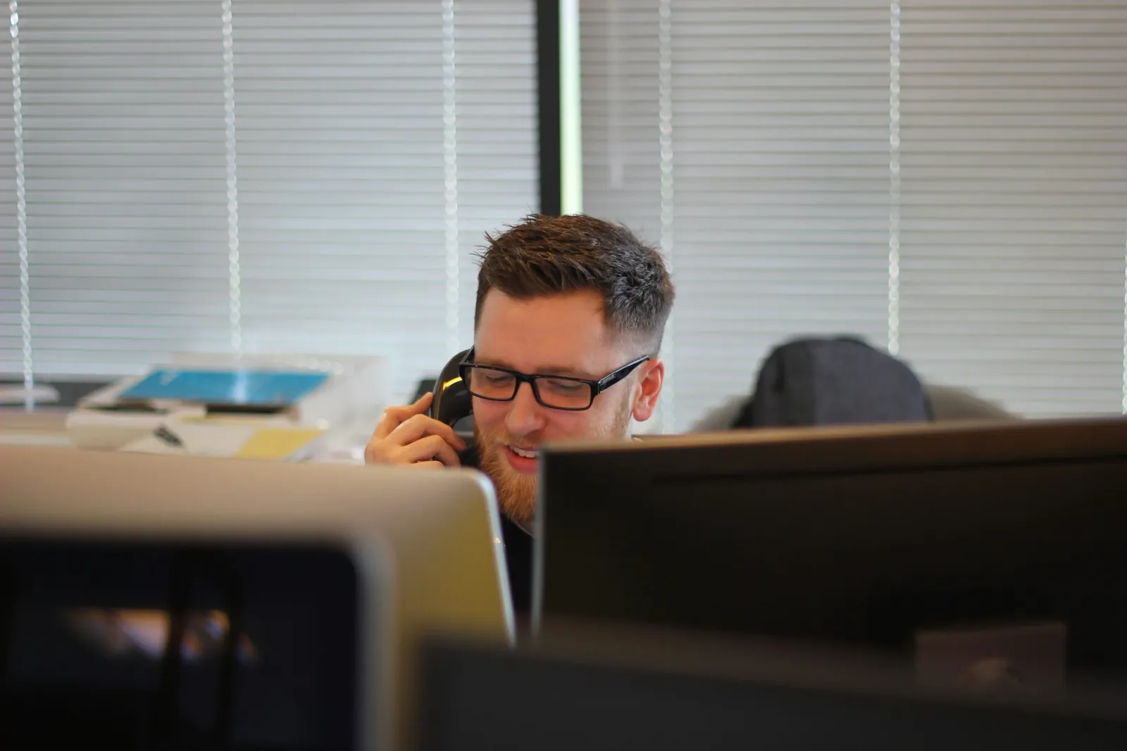 A man at an office desk taking a call on a corded desk phone, seen between two monitors.